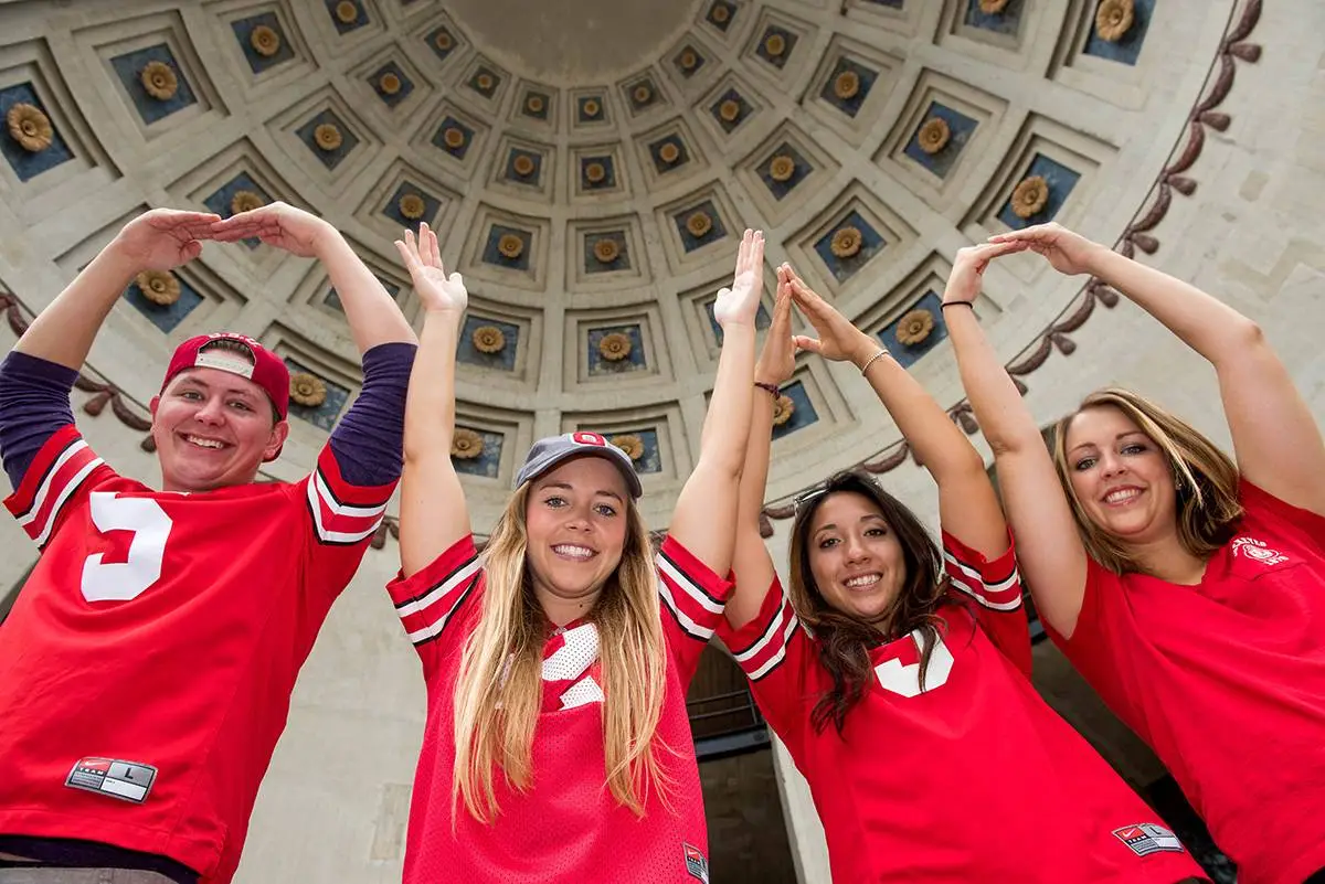 Four people wearing red shirts and caps pose with their arms forming the letters O-H-I-O. They stand under a dome with decorative patterns, smiling and looking joyful.