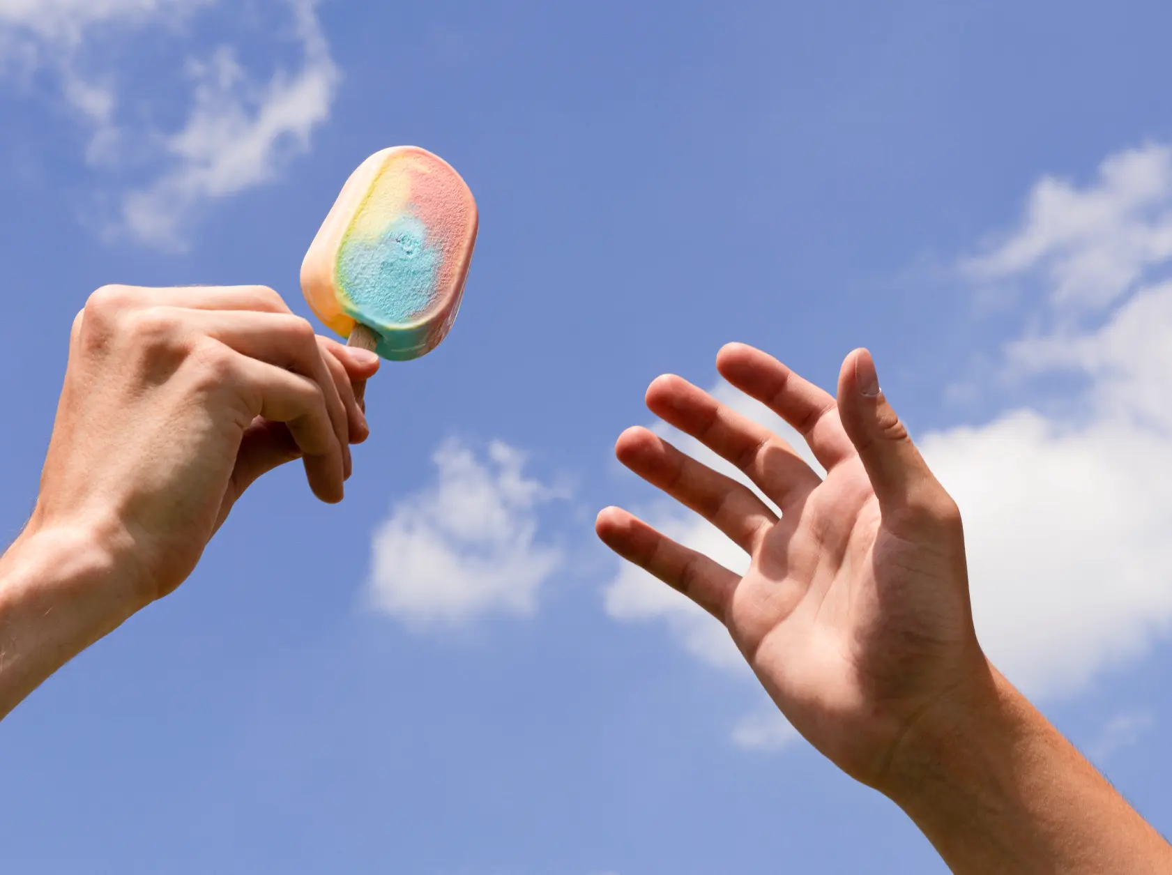A hand holds a colorful popsicle in the air while another hand reaches toward it, both set against a bright blue sky with a few scattered clouds.